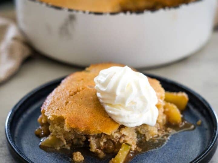 A slice of apple cobbler topped with whipped cream sits on a blue plate, with a larger dish of cobbler in the background.