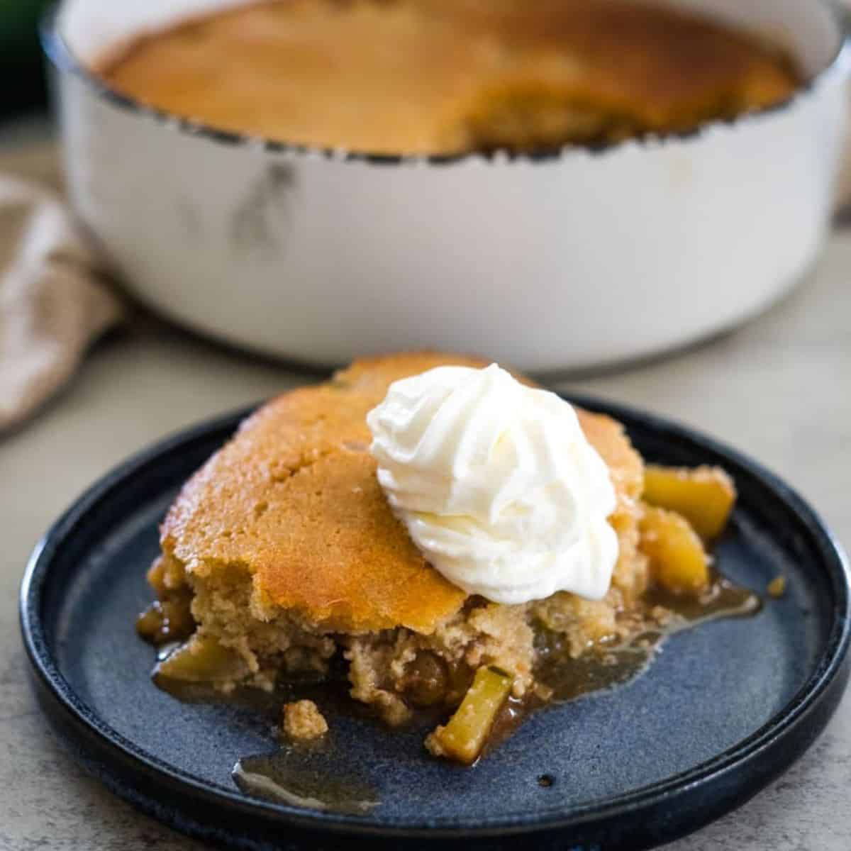 A slice of apple cobbler topped with whipped cream sits on a blue plate, with a larger dish of cobbler in the background.