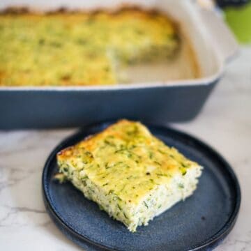 A piece of baked zucchini slice served on a black plate, with the remaining zucchini bake in a rectangular dish in the background.