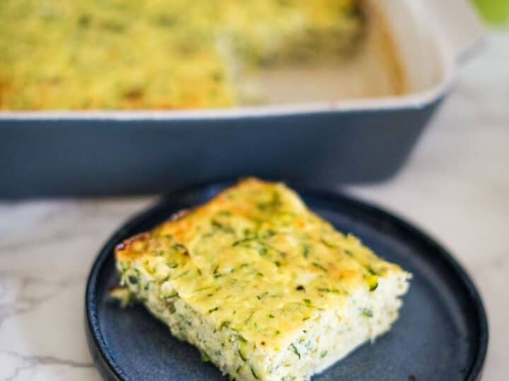 A piece of baked zucchini slice served on a black plate, with the remaining zucchini bake in a rectangular dish in the background.