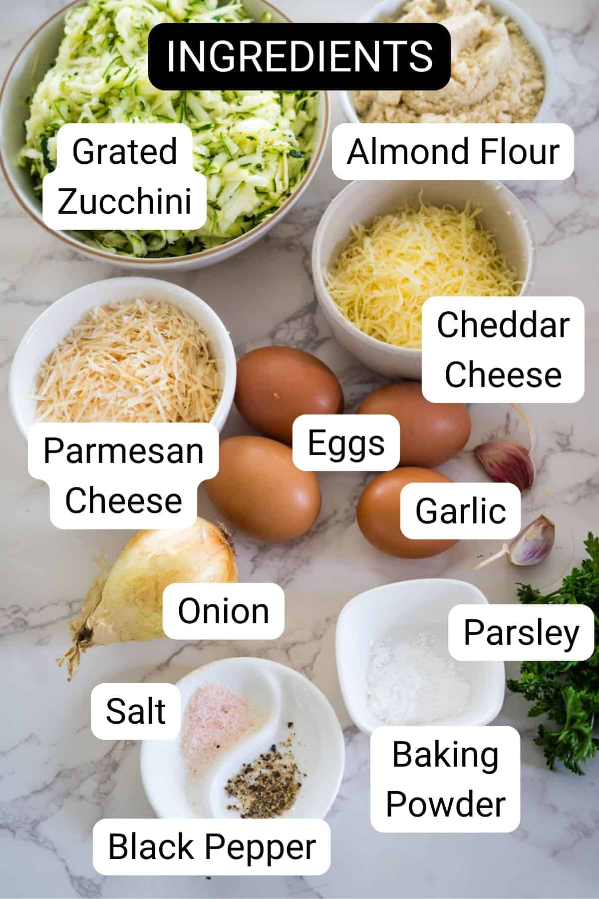 A marble counter displays labeled ingredients for a recipe: grated zucchini, almond flour, cheddar cheese, parmesan cheese, eggs, garlic, onion, parsley, salt, black pepper, and baking powder.