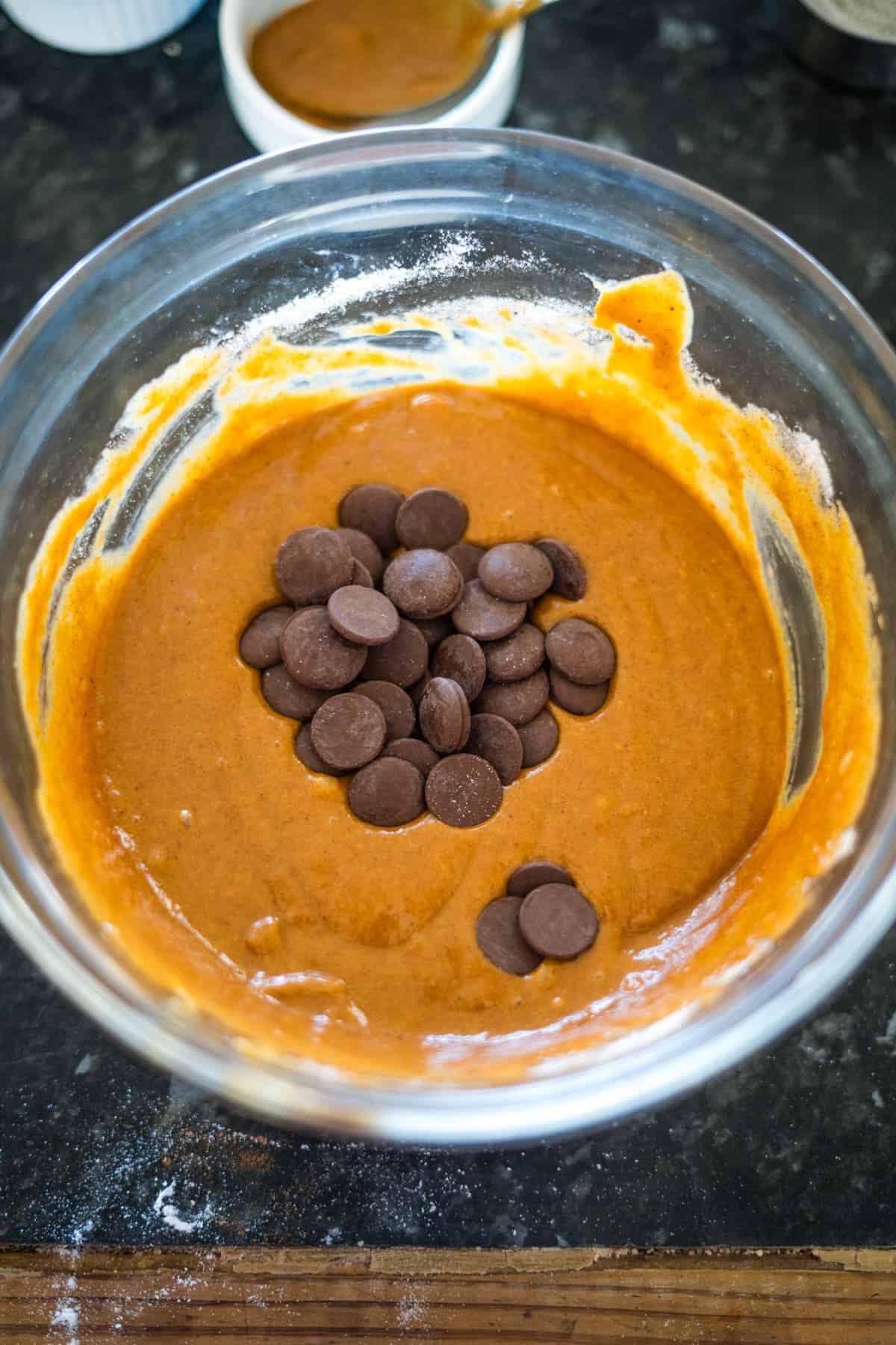 A glass bowl containing cookie batter with chocolate chips on top sits on a dark countertop, resembling the beginnings of flourless pumpkin chocolate chip muffins ready to be mixed in.