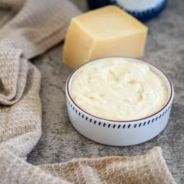 A small bowl of creamy white sauce next to a block of cheese and a beige textured cloth on a gray surface.