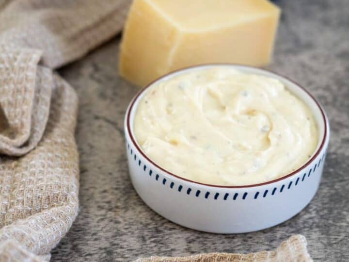 A small bowl of creamy white sauce next to a block of cheese and a beige textured cloth on a gray surface.