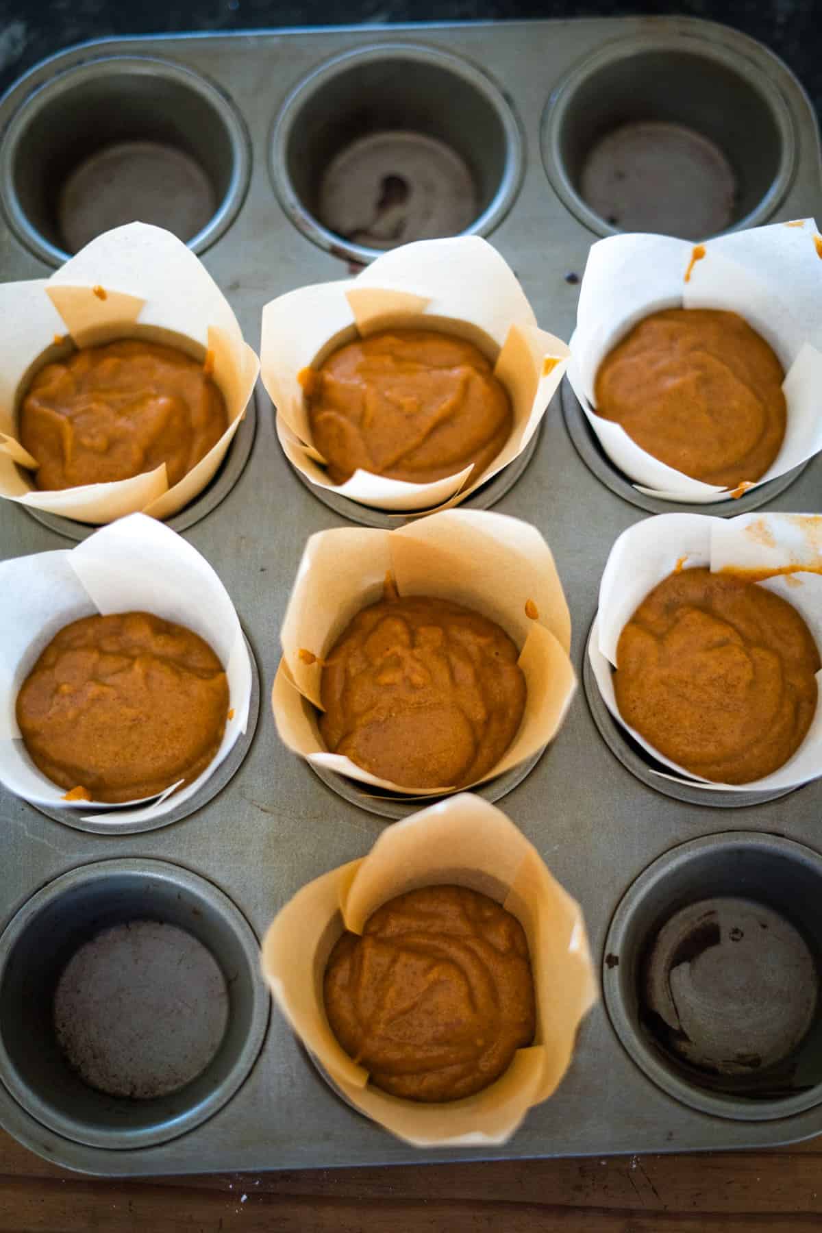 A muffin tin filled with six partially baked flourless pumpkin chocolate chip muffins in parchment paper cups, with two empty muffin slots.