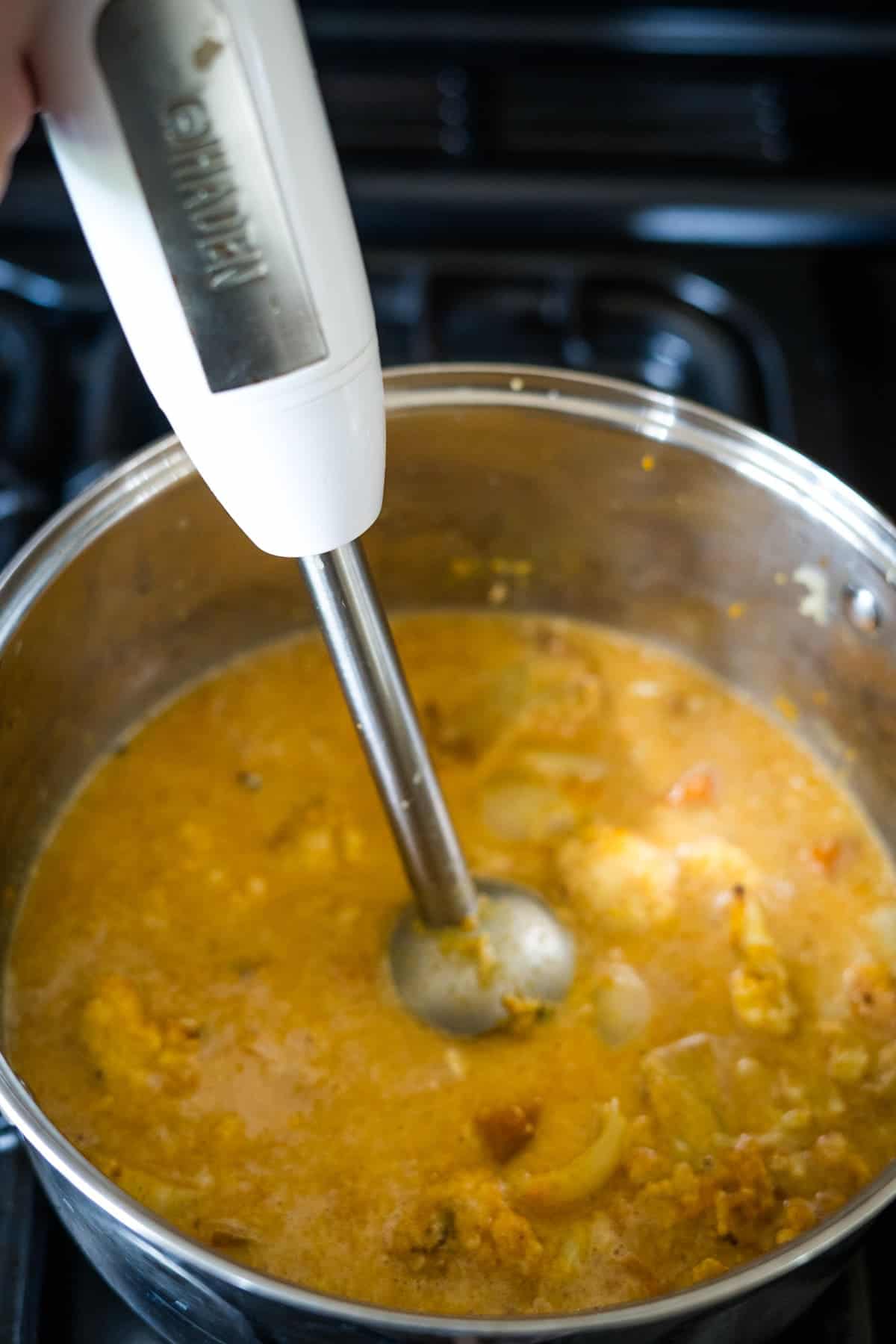 A hand uses an immersion blender in a pot of pumpkin cauliflower soup, blending the orange mixture that contains chunks of vegetables.