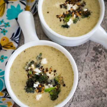 Two bowls of creamy soup garnished with broccoli, cheese, and seasoning on a textured surface, alongside a floral-patterned napkin.