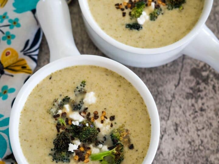 Two bowls of creamy soup garnished with broccoli, cheese, and seasoning on a textured surface, alongside a floral-patterned napkin.