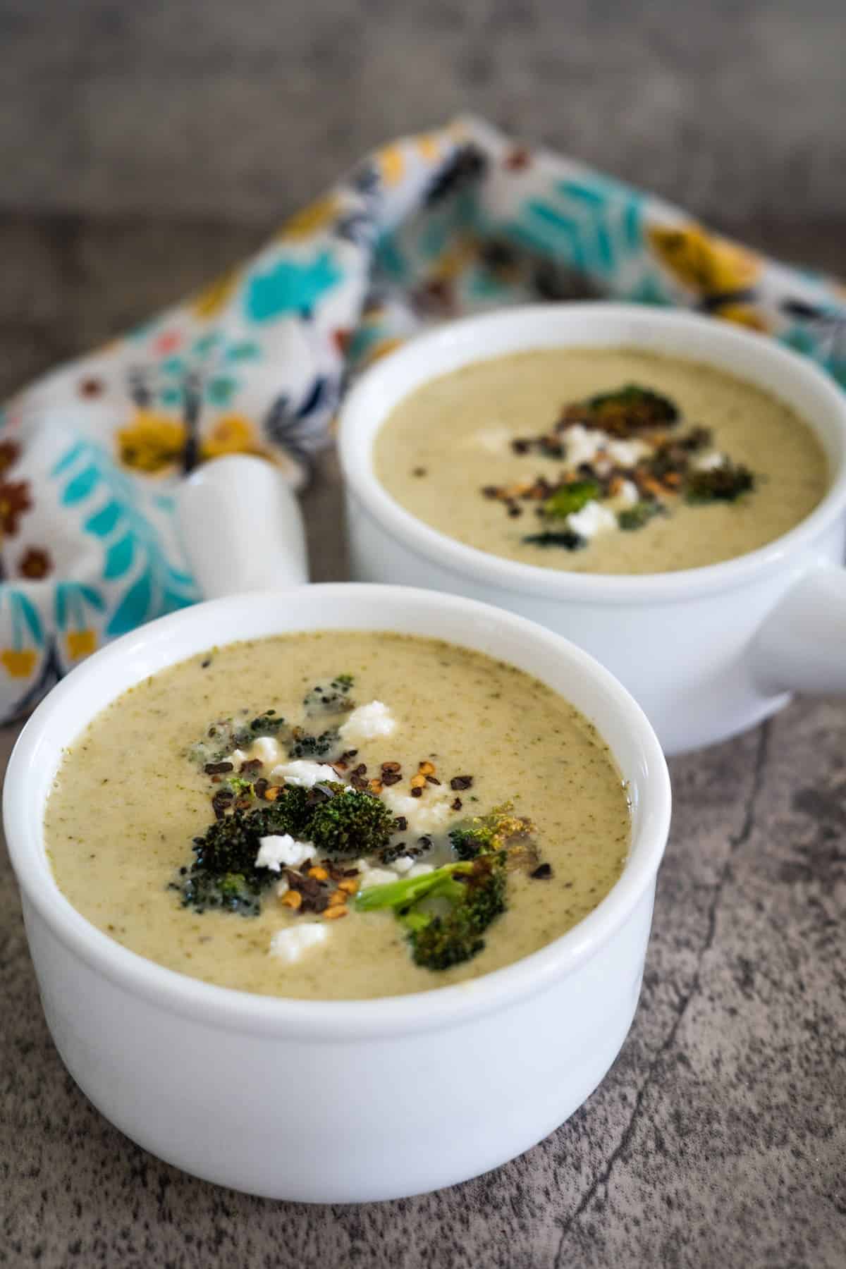 Two white bowls filled with creamy broccoli soup topped with roasted broccoli florets and a sprinkle of spices. A colorful patterned napkin is in the background.