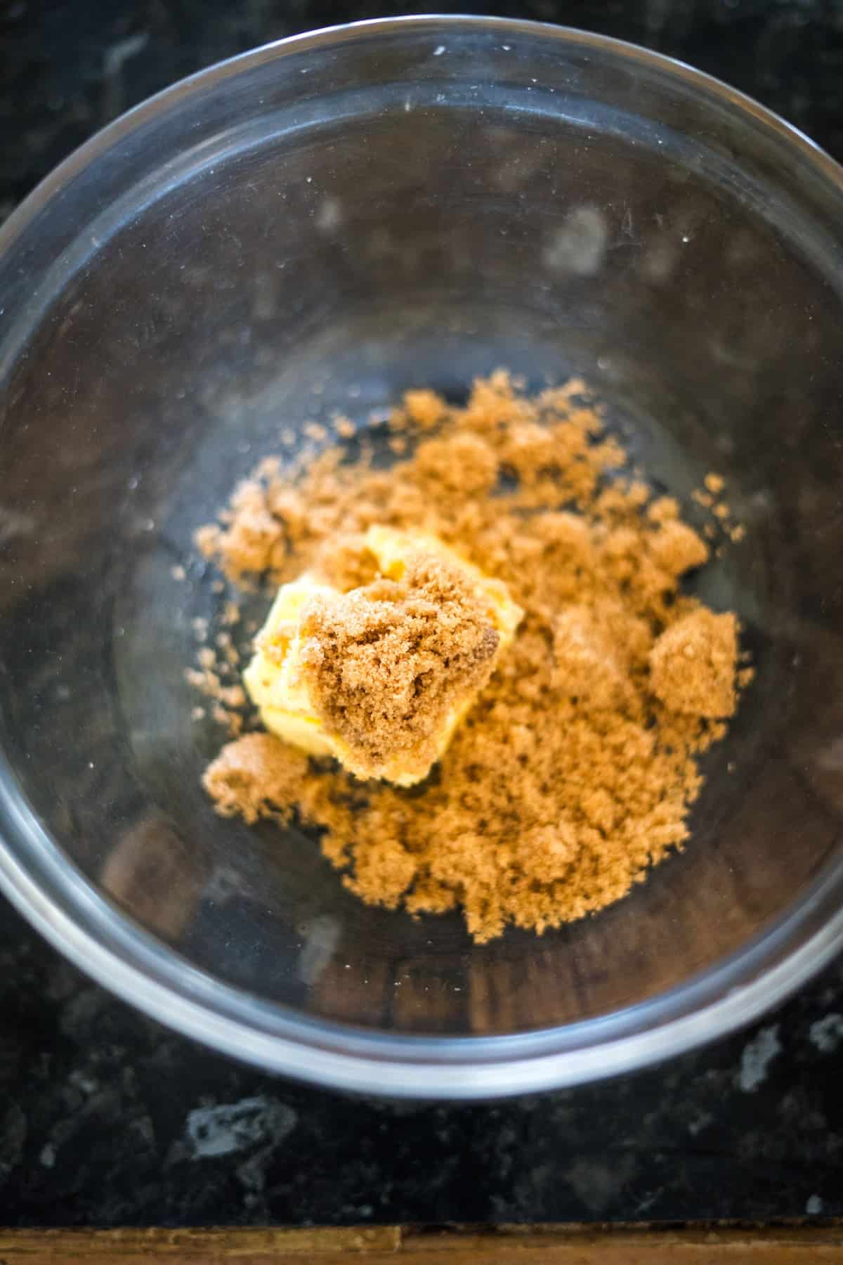 A glass bowl contains a mixture of butter and brown sugar on a dark counter, perfect for making keto pumpkin cheesecake muffins.