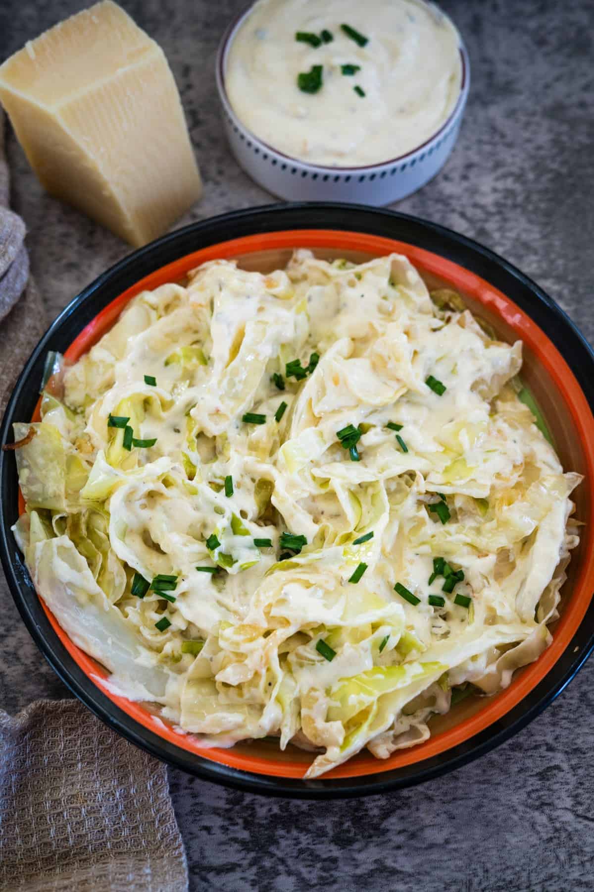 A colorful bowl of cabbage salad topped with dressing and chopped chives, positioned next to a block of cheese and a small dish of additional dressing.