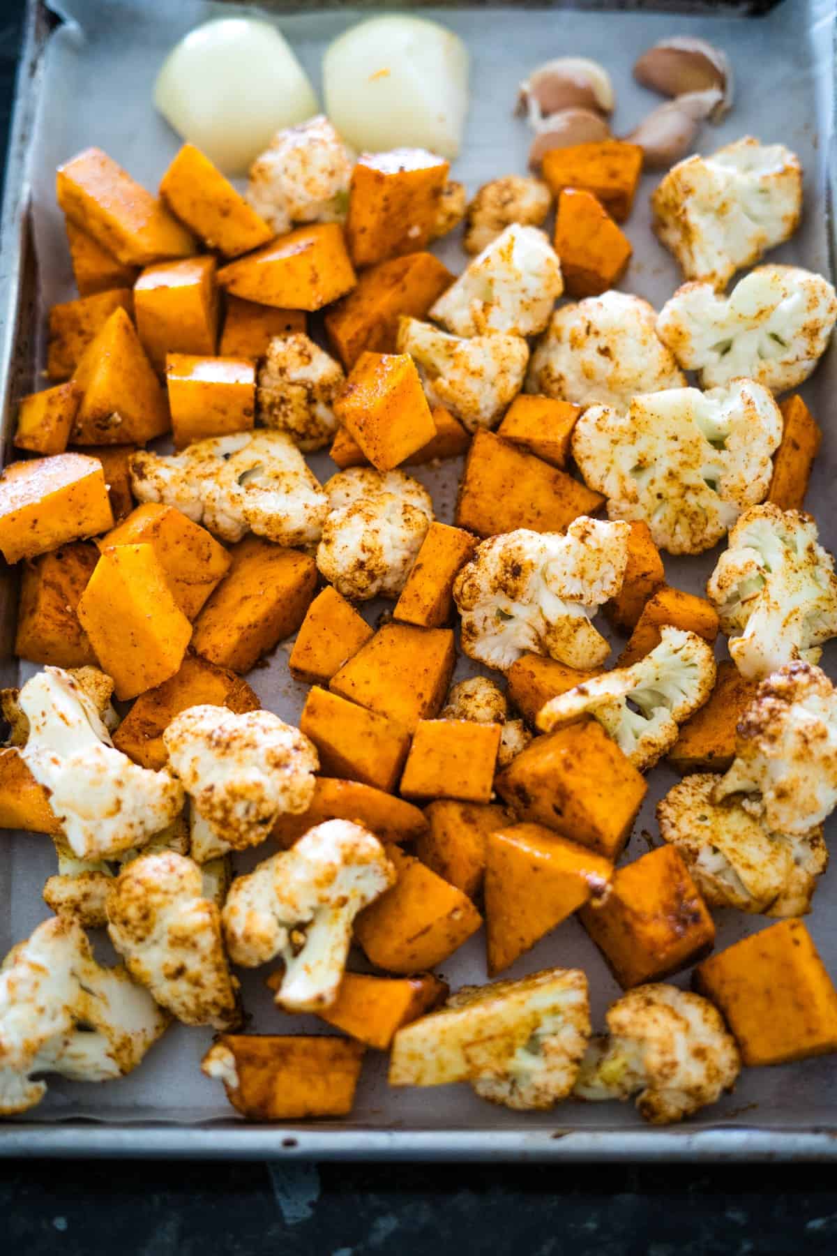 A baking sheet with cubed sweet potatoes, cauliflower florets, whole garlic cloves, and halved onions, all seasoned and ready for roasting—perfect ingredients to add depth to a pumpkin cauliflower soup.