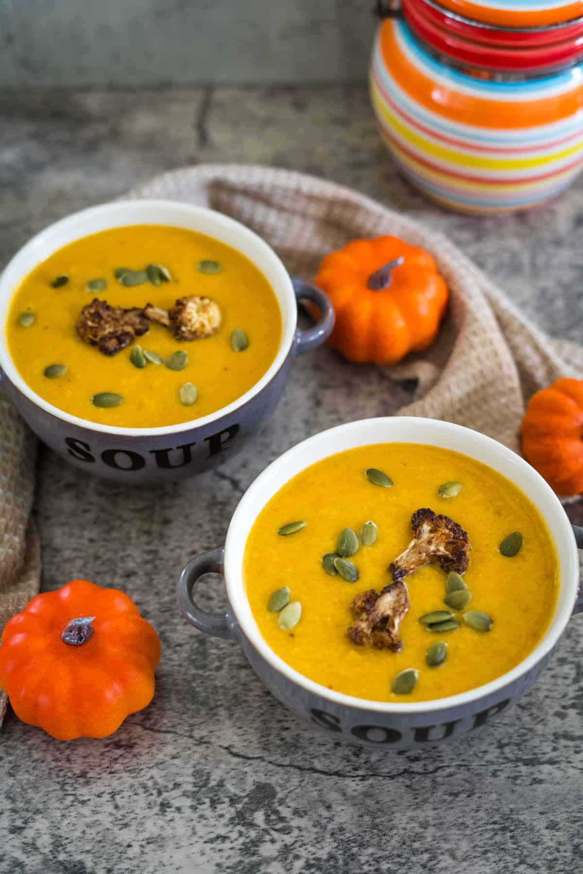 Two bowls of pumpkin cauliflower soup garnished with roasted seeds, surrounded by small pumpkins and a textured fabric on a stone surface.