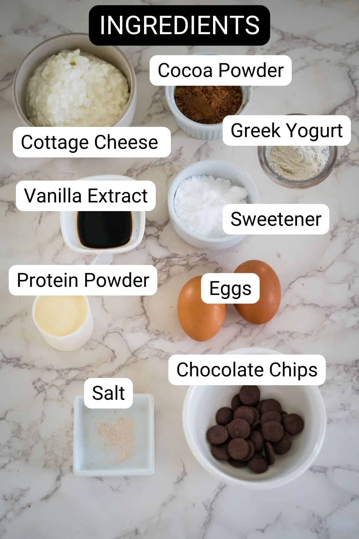 A marble countertop displays labeled bowls containing cottage cheese, cocoa powder, Greek yogurt, vanilla extract, sweetener, protein powder, eggs, salt, and chocolate chips.