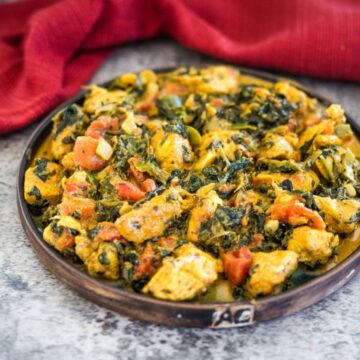 A plate of chicken and vegetable curry with chunks of chicken, leafy greens, and diced tomatoes, garnished with herbs. A red cloth is in the background.