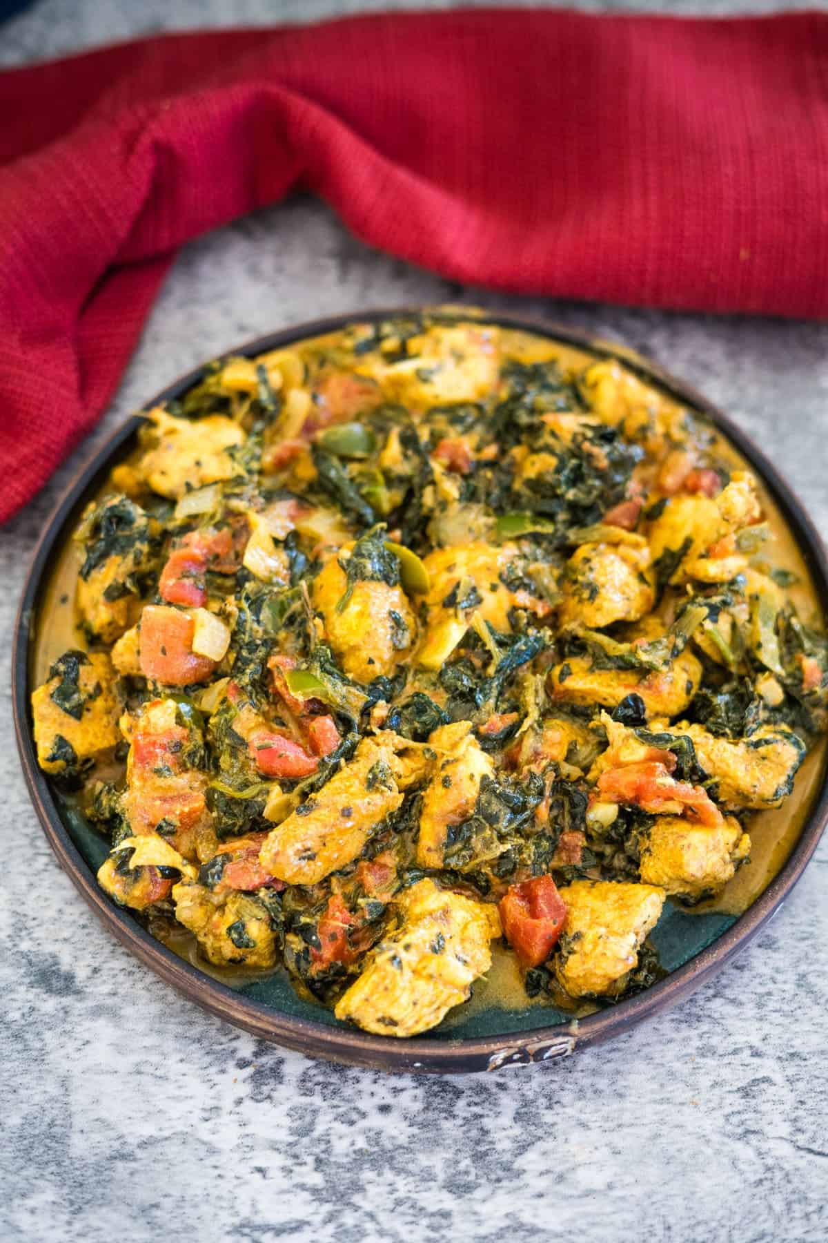 A plate of palak chicken featuring cooked chicken pieces mixed with spinach and diced tomatoes, set on a gray surface with a red cloth in the background.