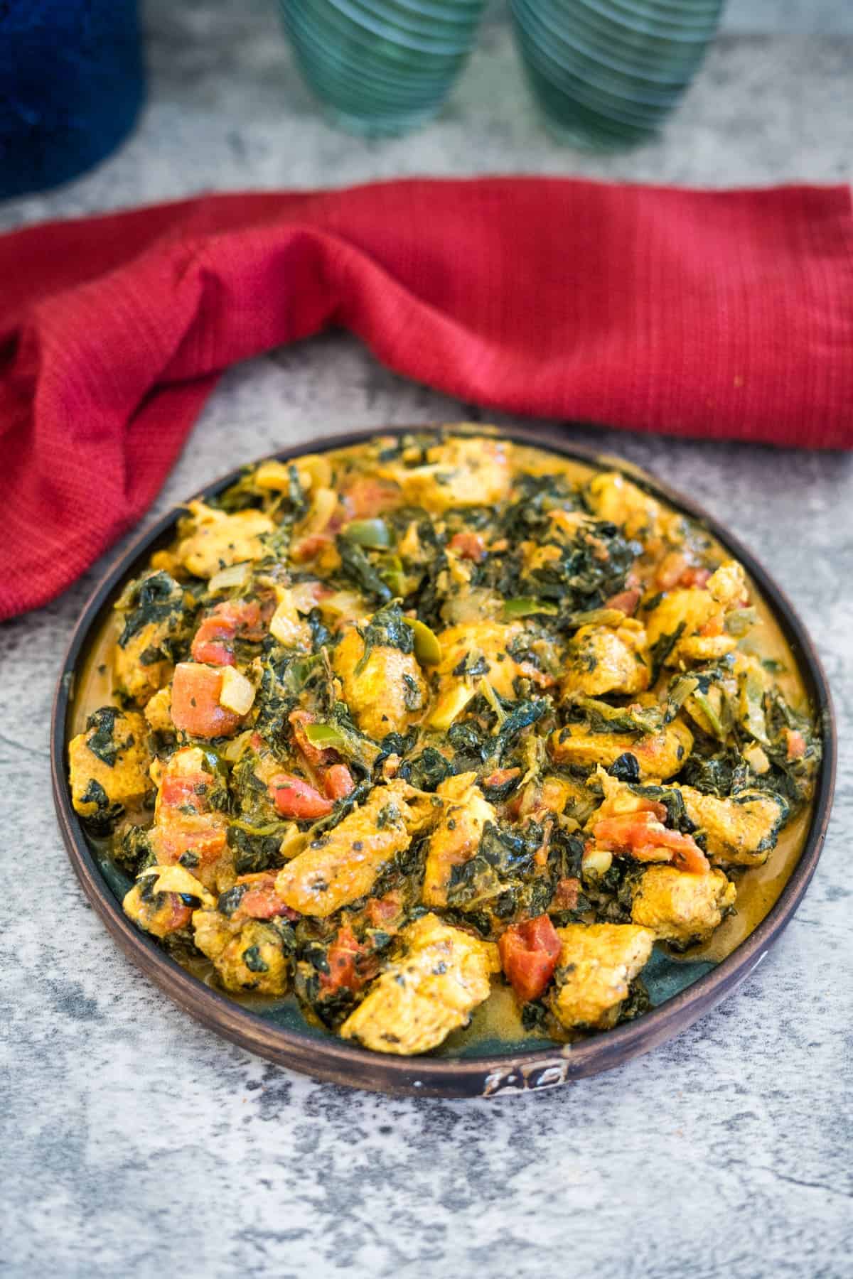 A plate of palak chicken, featuring tender cooked pieces mixed with spinach, tomatoes, and spices, is placed on a grey surface with a red cloth casually positioned behind it.