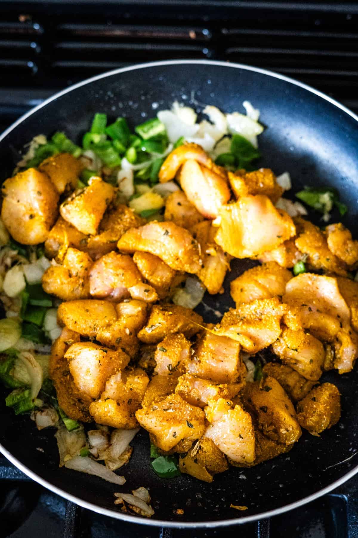 Chunks of seasoned chicken cooking with chopped onions and green bell peppers in a black skillet on a stove.