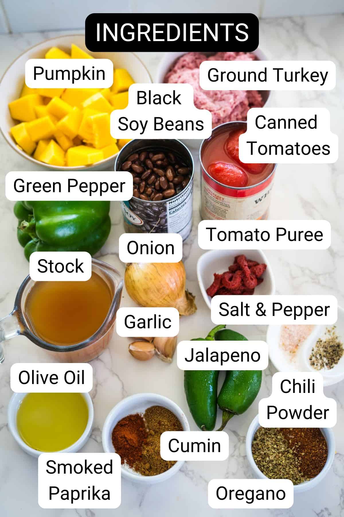 Image showing various labeled ingredients on a kitchen counter, including pumpkin, ground turkey, black soy beans, canned tomatoes, green pepper, stock, onion, tomato puree, garlic, and more for a delicious keto pumpkin chili.