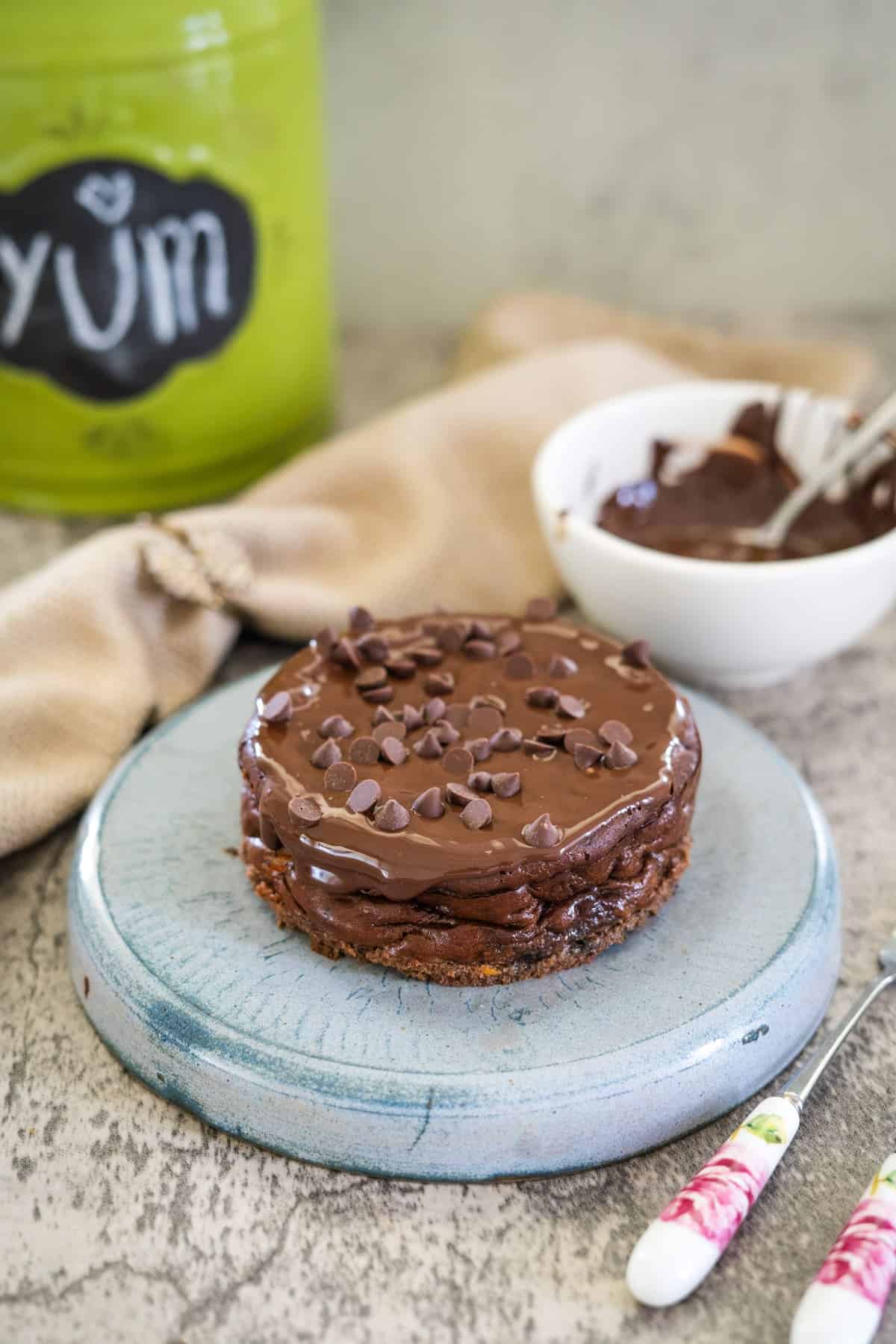 A chocolate cake topped with rich chocolate frosting and mini chocolate chips on a blue plate, with a bowl of frosting and utensils in the background, evokes the indulgence of a chocolate cottage cheesecake delight.