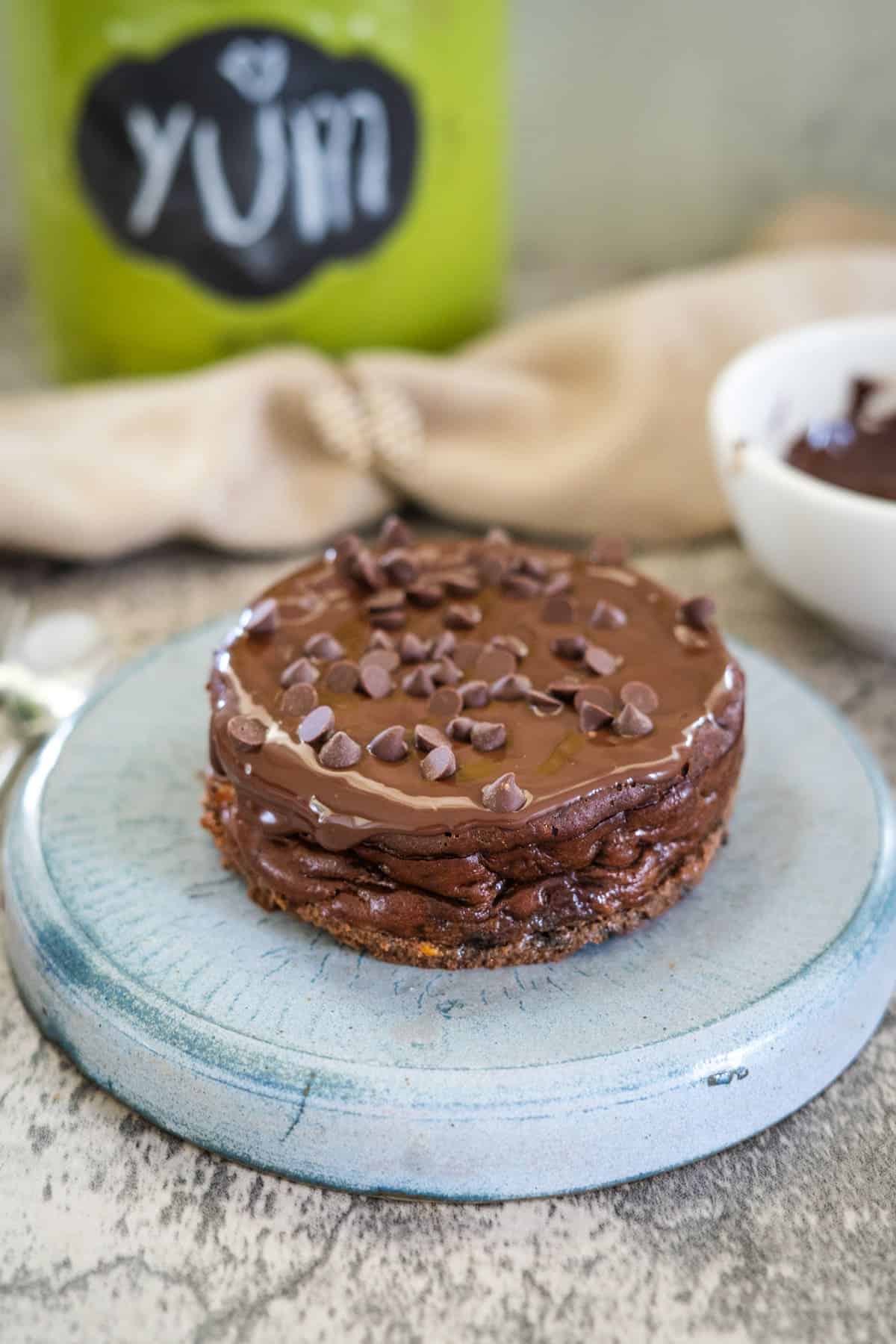 A chocolate dessert topped with small chocolate chips on a blue ceramic plate. A white bowl and a container labeled "yum" are in the background.