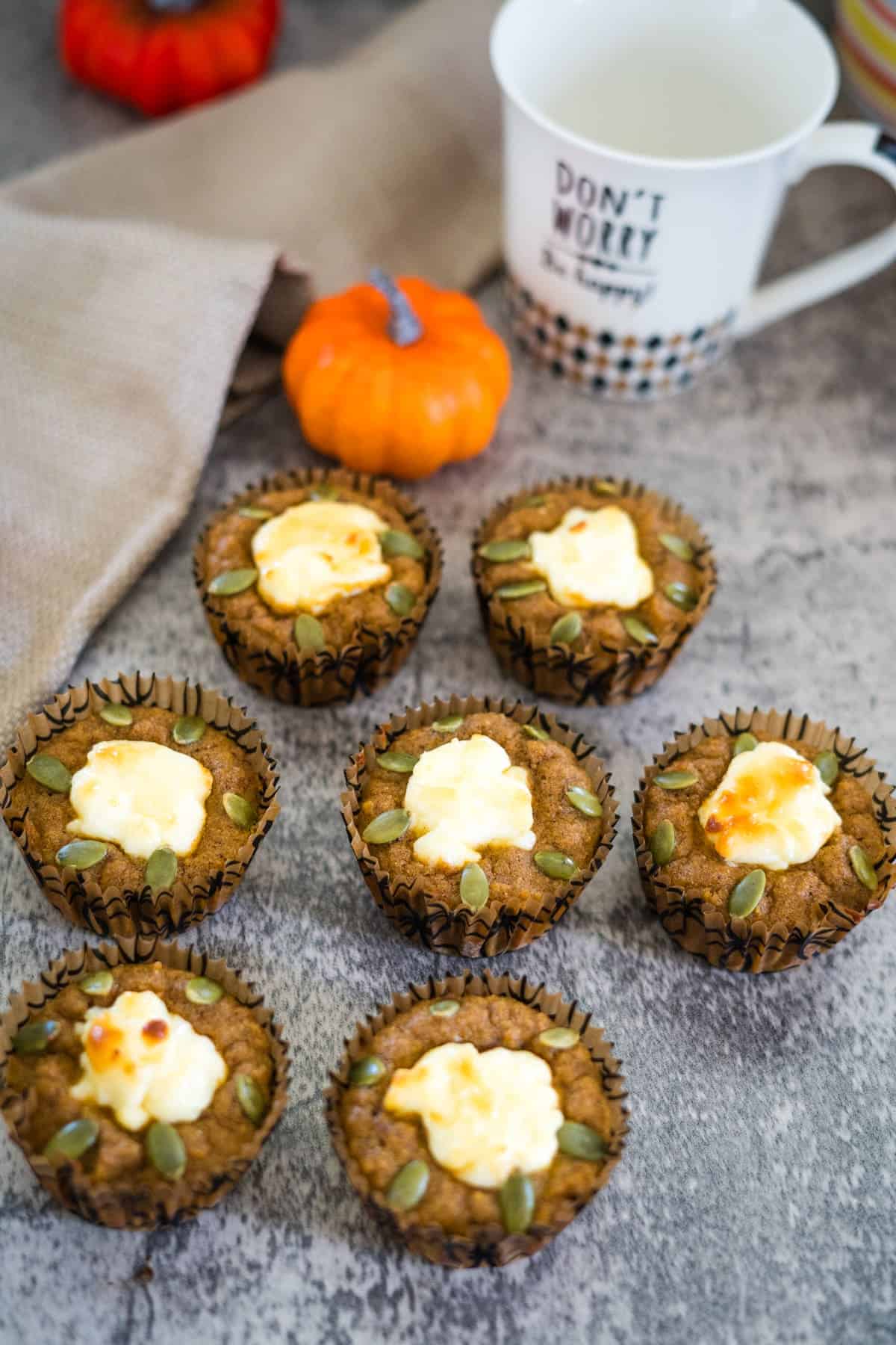 Six keto pumpkin cheesecake muffins with cream cheese and pumpkin seed toppings arranged on a gray surface near a beige napkin, small decorative pumpkins, and a white mug with text.