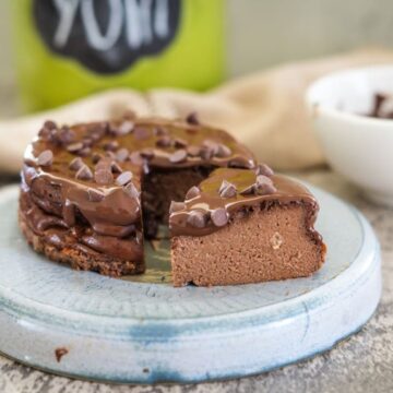 A chocolate cake with a slice removed sits on a blue plate. The cake is topped with chocolate frosting and chocolate chips. A bowl and a jar labeled "YUM" are blurred in the background.