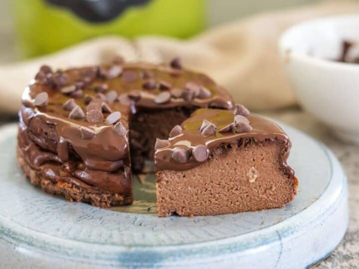 A chocolate cake with a slice removed sits on a blue plate. The cake is topped with chocolate frosting and chocolate chips. A bowl and a jar labeled "YUM" are blurred in the background.