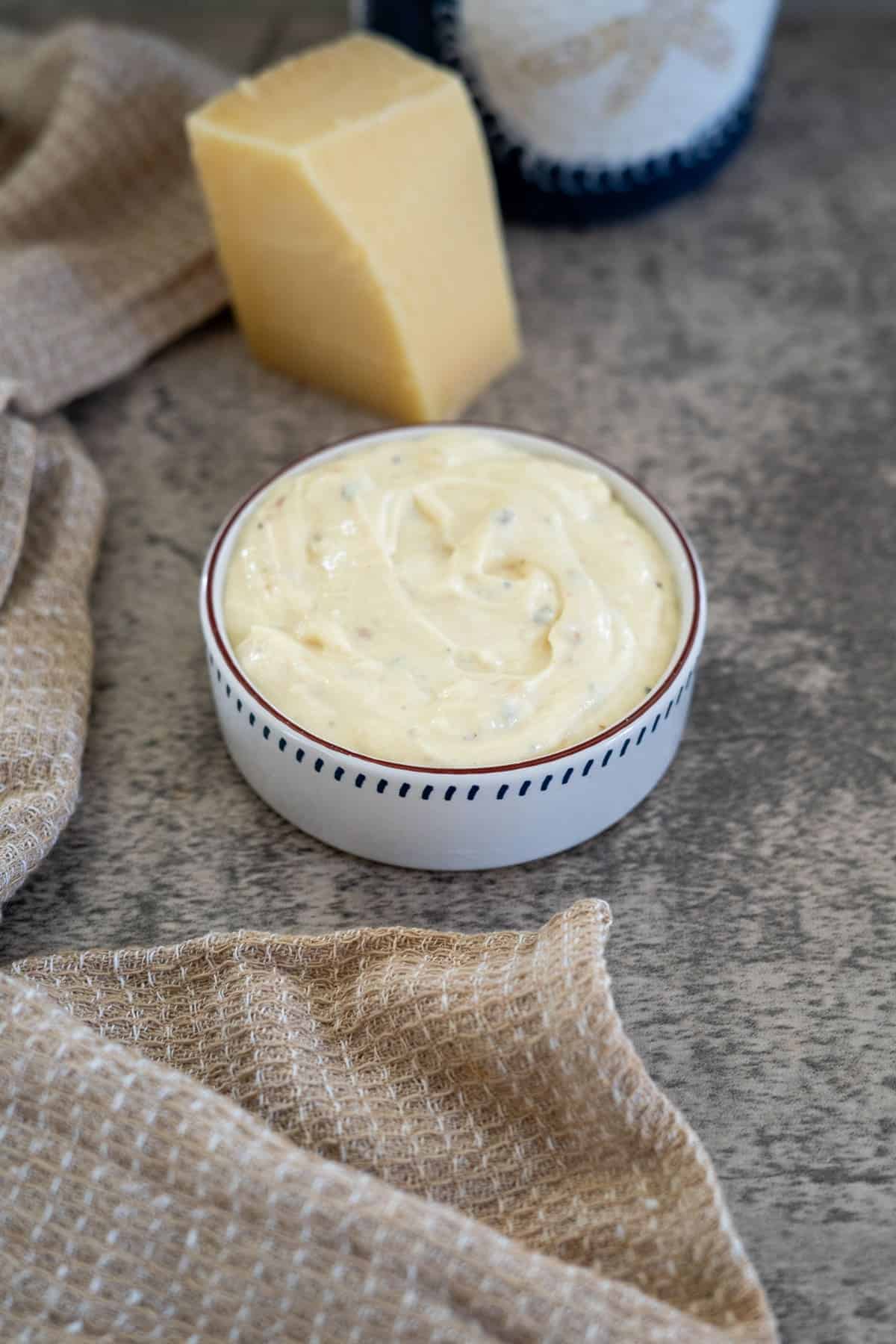 A small bowl filled with creamy dip is placed next to a wedge of cheese and a beige cloth on a gray surface.