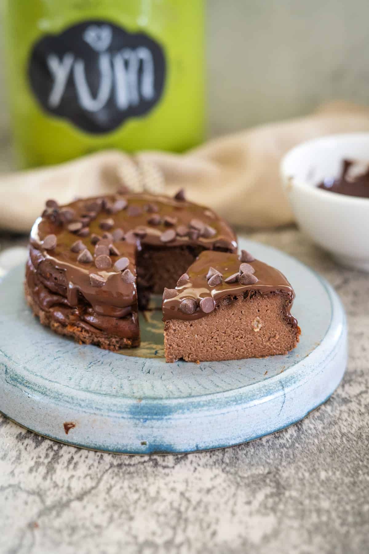 A small, round chocolate cottage cheese cheesecake on a blue ceramic plate with a slice cut out. The cheesecake is topped with chocolate chips and there is a green jar with "YUM" written on it in the background.