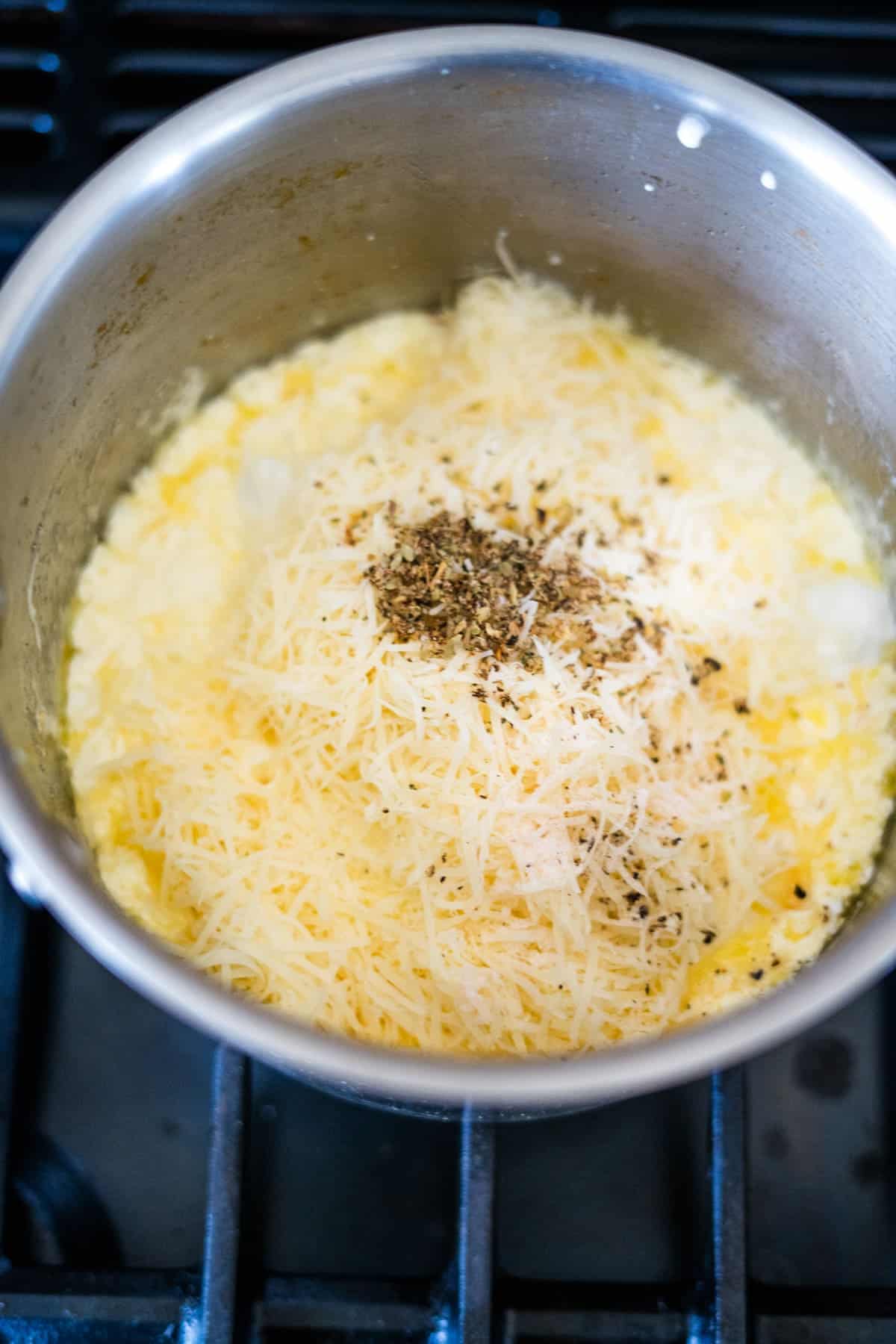 A stainless steel pot on a stove with scrambled eggs, grated cheese, and black pepper on top.