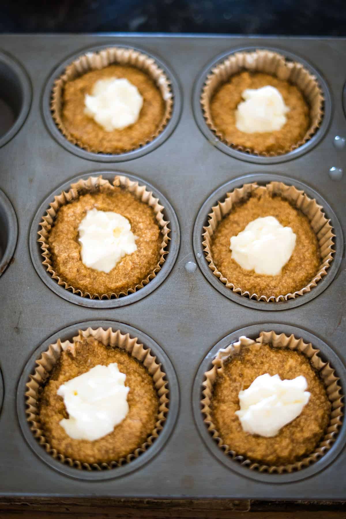 Six keto pumpkin cheesecake muffin batter portions in a muffin tin, each topped with a dollop of white filling, ready to be baked.