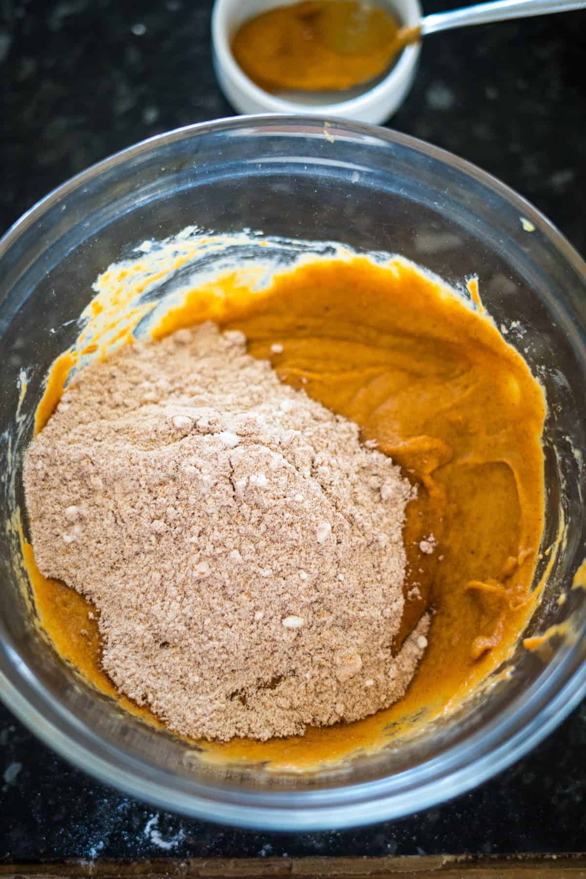 A clear mixing bowl contains a partially mixed mixture of orange batter and a pile of dry ingredients, likely for flourless pumpkin chocolate chip muffins. A white bowl with a spoon rests in the background on a black countertop.