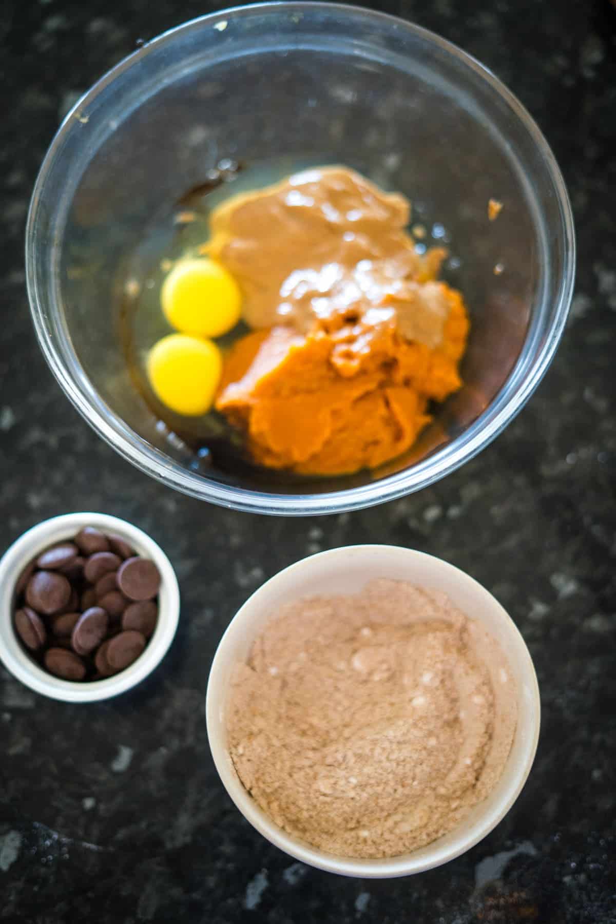 A mixing bowl with eggs, peanut butter, and sugar, alongside bowls of flour mixture and chocolate chips on a dark countertop, ready to create flourless pumpkin chocolate chip muffins.