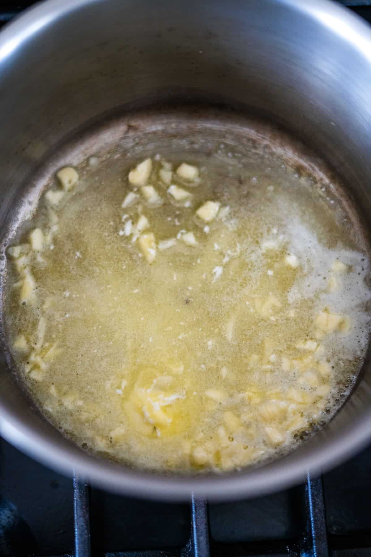 A stainless steel saucepan on a stovetop with melted butter and minced garlic being sautéed.