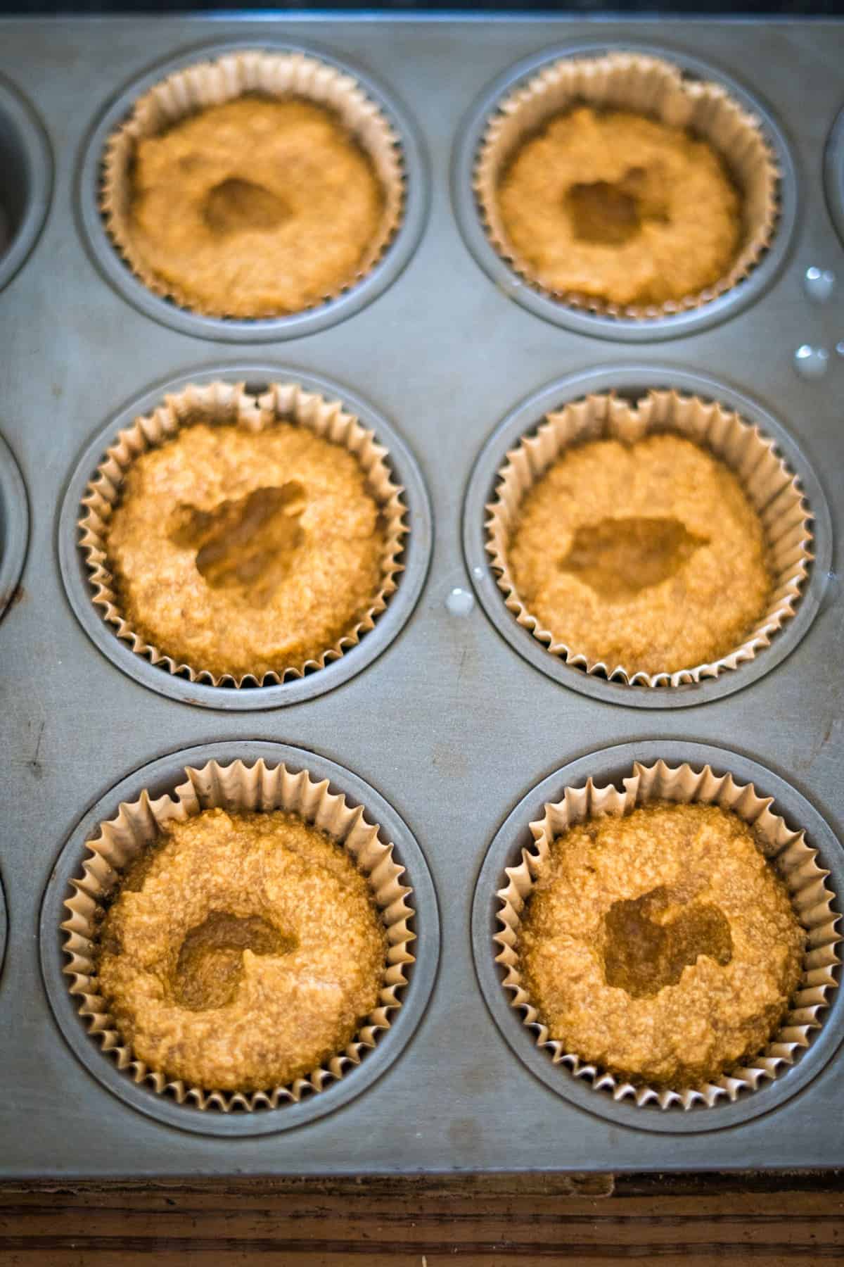 A muffin tray filled with six portions of uncooked keto pumpkin cheesecake muffin batter in paper liners, ready for baking.