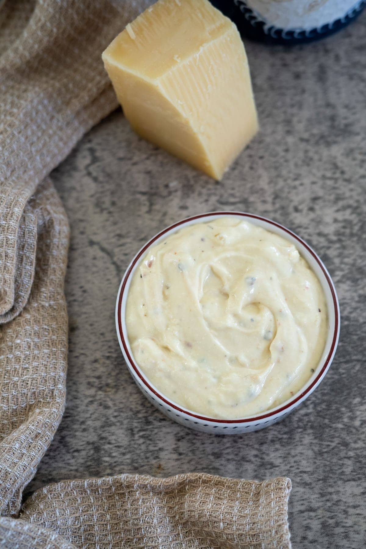 A small bowl of creamy dip is placed on a textured surface next to a block of cheese and a folded beige cloth.