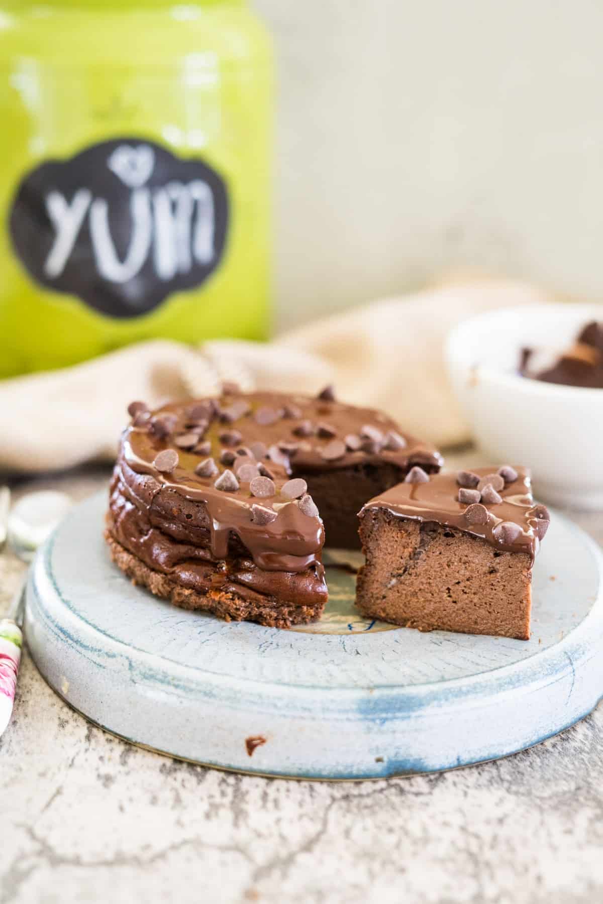 A chocolate cake topped with chocolate chips on a plate, with one slice cut and slightly separated from the rest. A green jar labeled "YUM" is in the background, adding a touch of charm to this tempting dessert reminiscent of a rich chocolate cheesecake.