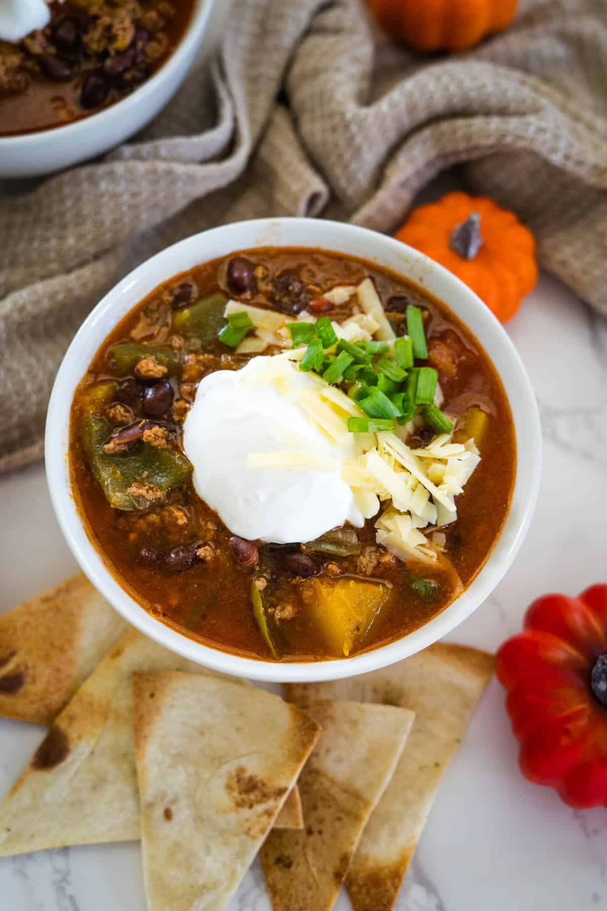 A bowl of keto pumpkin chili topped with sour cream, shredded cheese, and chopped green onions, surrounded by tortilla chips and small decorative pumpkins.