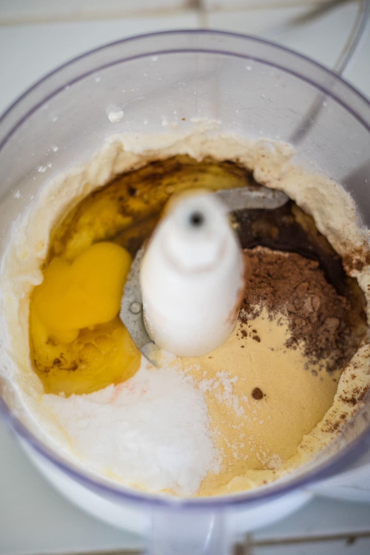 A food processor container with eggs, flour, cocoa powder, sugar, and other ingredients ready to be mixed for a chocolate cheesecake.