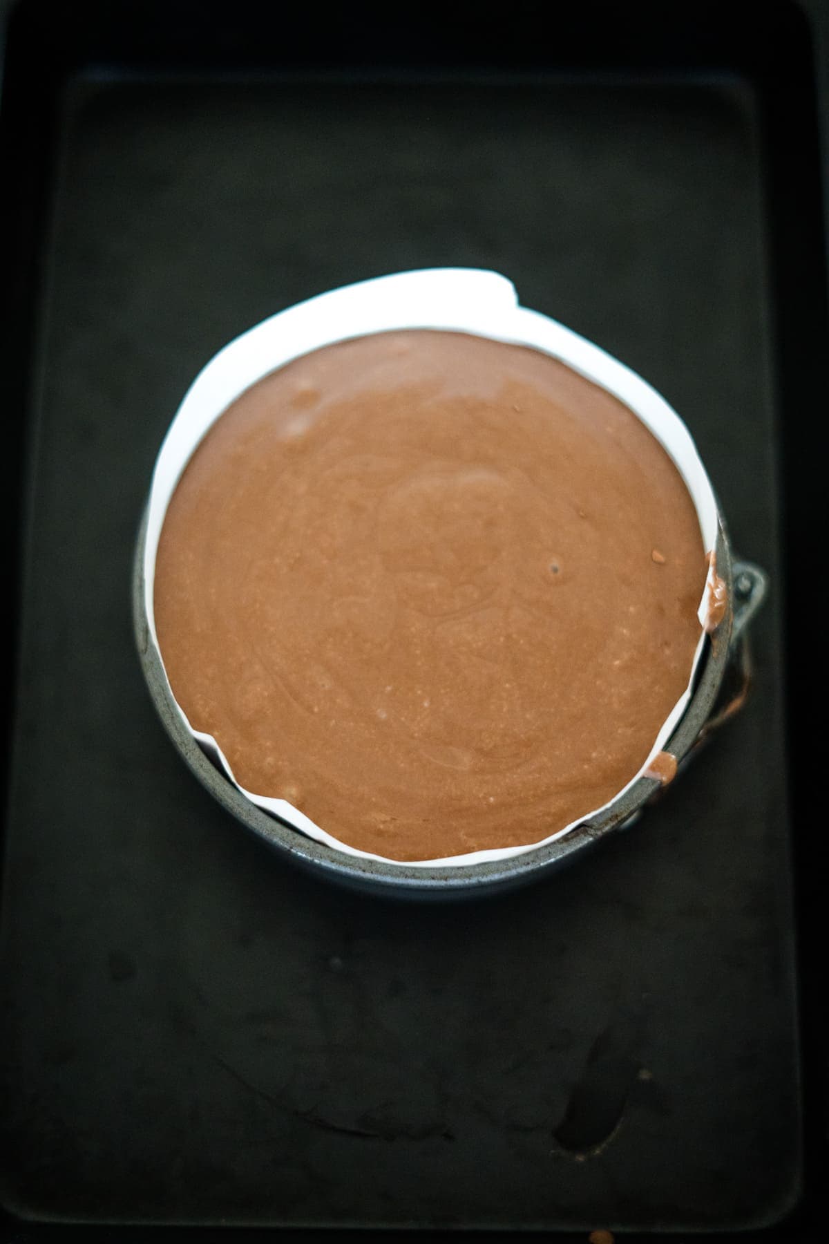 A round chocolate cake batter in a baking pan placed on a dark surface, ready to be baked.