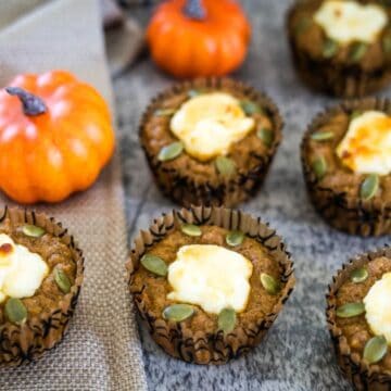 Pumpkin muffins with cream cheese centers and pumpkin seed toppings, placed on a grey surface with small decorative pumpkins in the background.