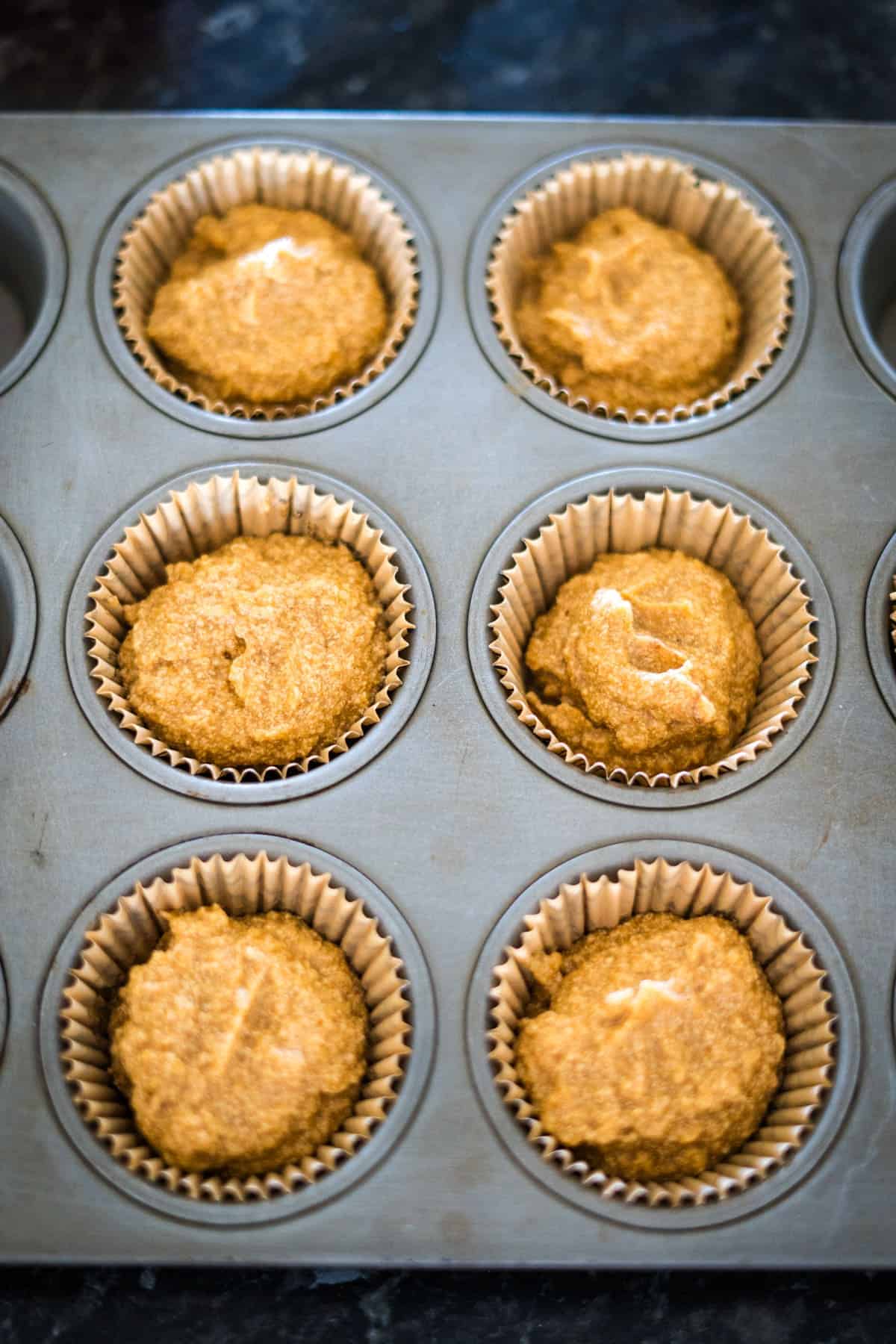 A close-up of a muffin pan with six paper liners, each filled with raw keto pumpkin cheesecake muffin batter, ready to be baked.