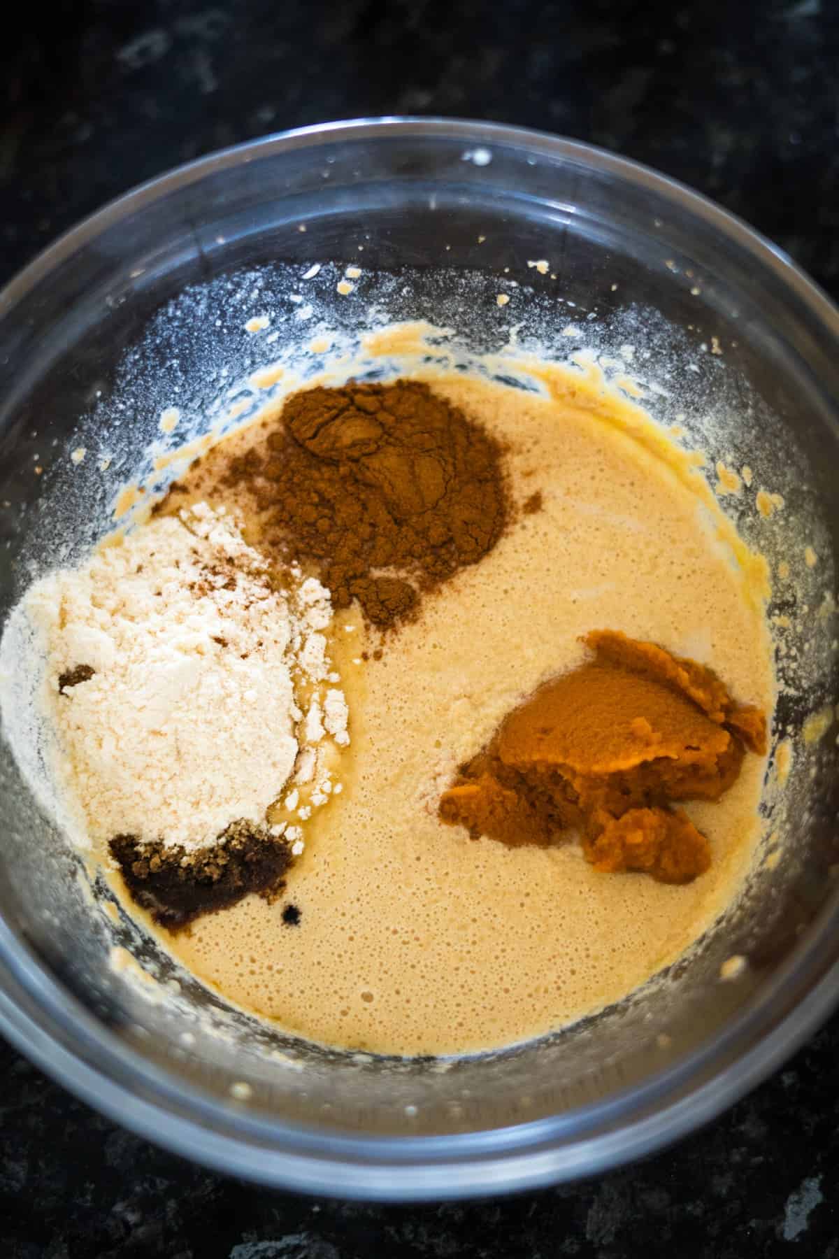 A glass bowl containing a beige batter mixed with flour, cinnamon, and brown sugar for keto pumpkin cheesecake muffins, set on a dark countertop.