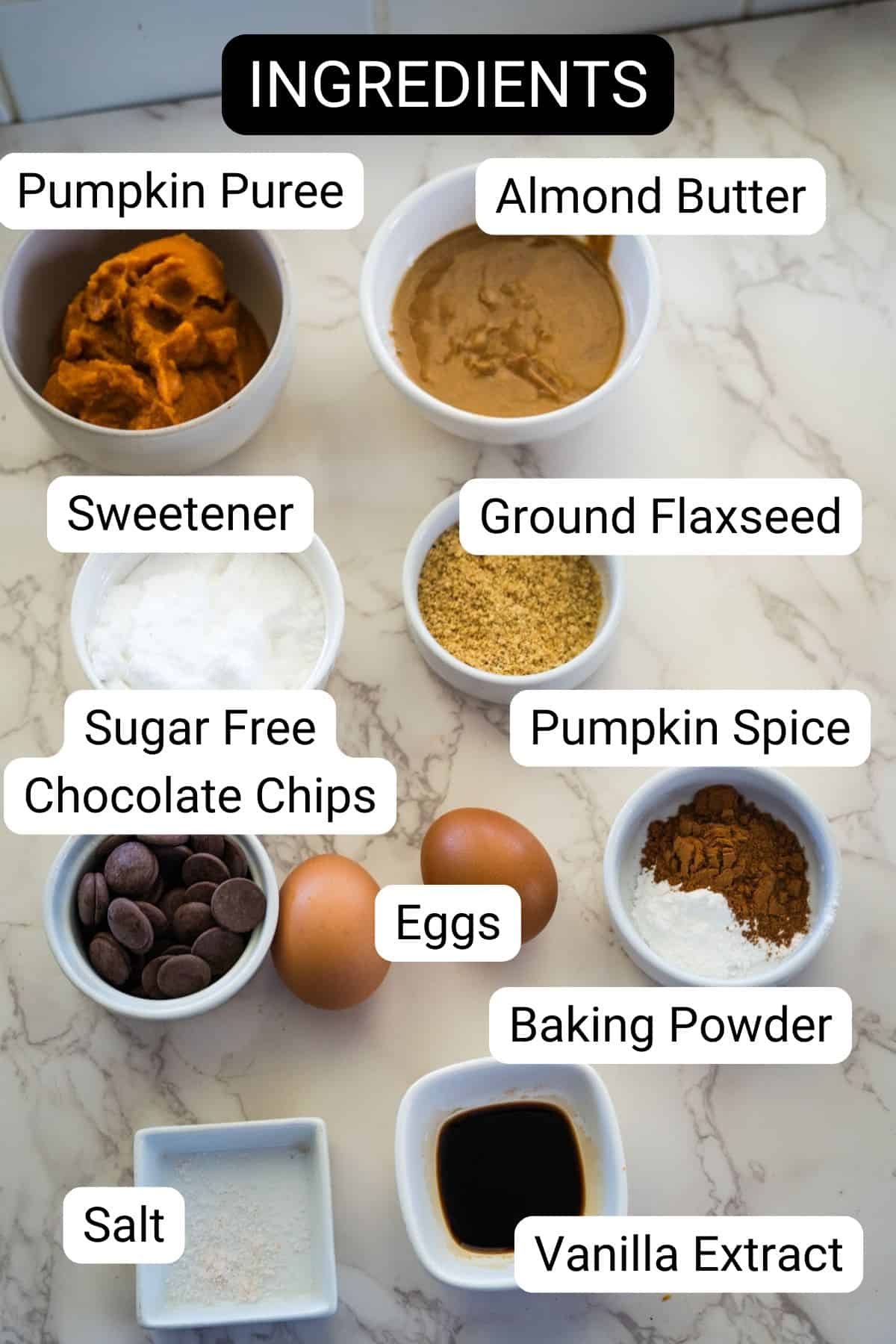 A marble countertop with measured ingredients in bowls: pumpkin puree, almond butter, sweetener, ground flaxseed, sugar-free chocolate chips, pumpkin spice, eggs, baking powder, salt, and vanilla extract.