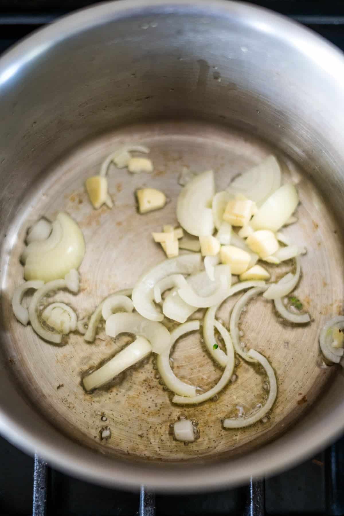 A stainless steel pot on a stove with sautéing onions and garlic slices.