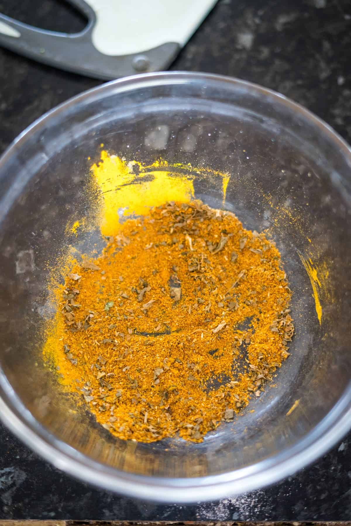 A clear glass bowl containing a mixture of ground spices, primarily yellow and brown in color, sits on a dark speckled countertop. A kitchen utensil is partially visible in the background.