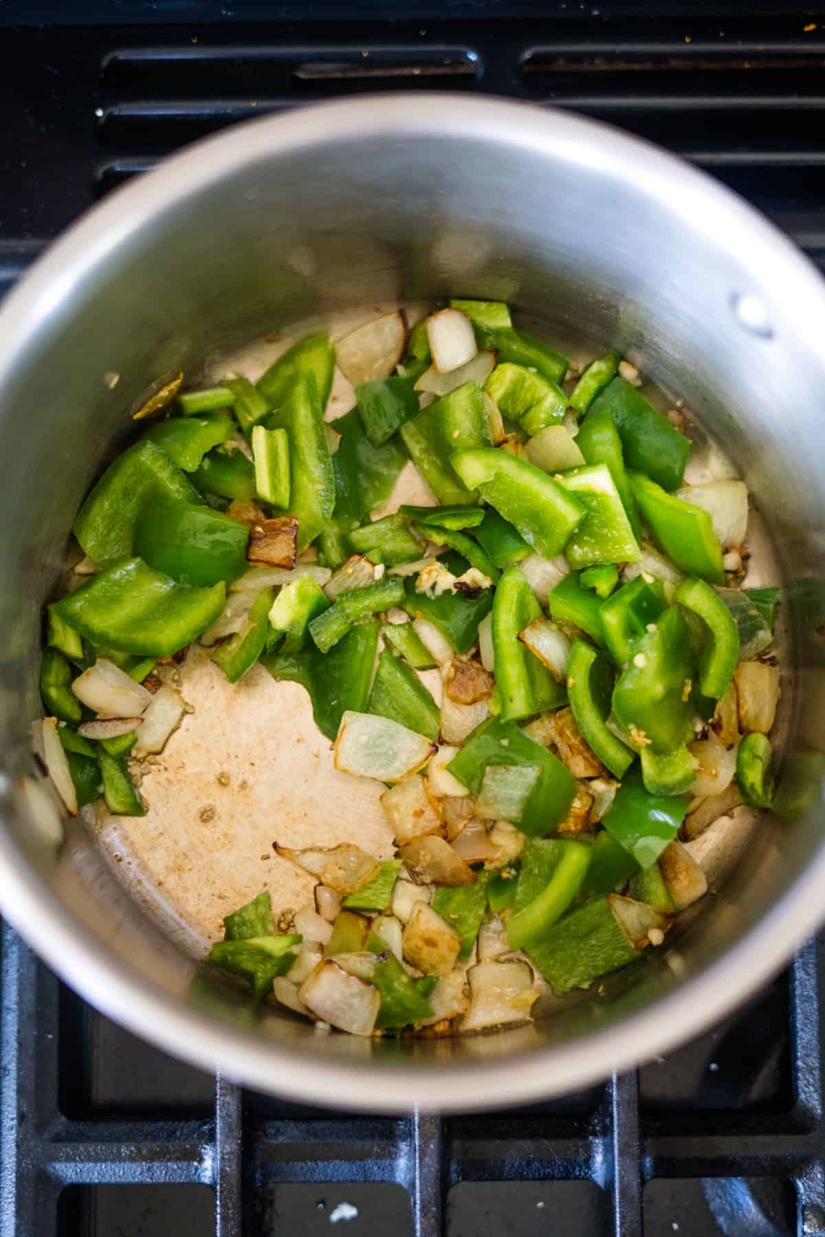 A stainless steel pot containing chopped green bell peppers and onions being sautéed on a stove, perfect for starting a delicious keto pumpkin chili.