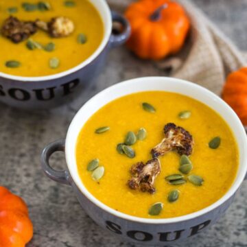 Two bowls of pumpkin soup garnished with roasted cauliflower and pumpkin seeds, surrounded by small decorative pumpkins.