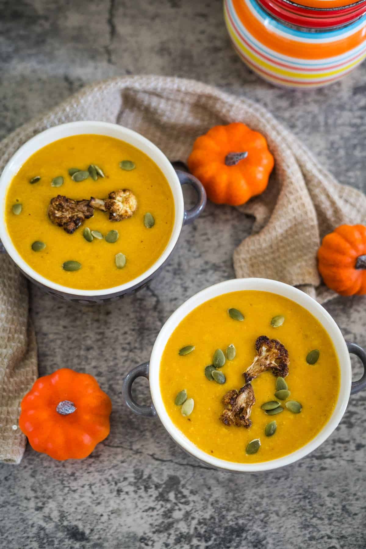 Two bowls of pumpkin cauliflower soup garnished with roasted cauliflower and pumpkin seeds, placed on a textured surface with small decorative pumpkins and a light brown cloth nearby.